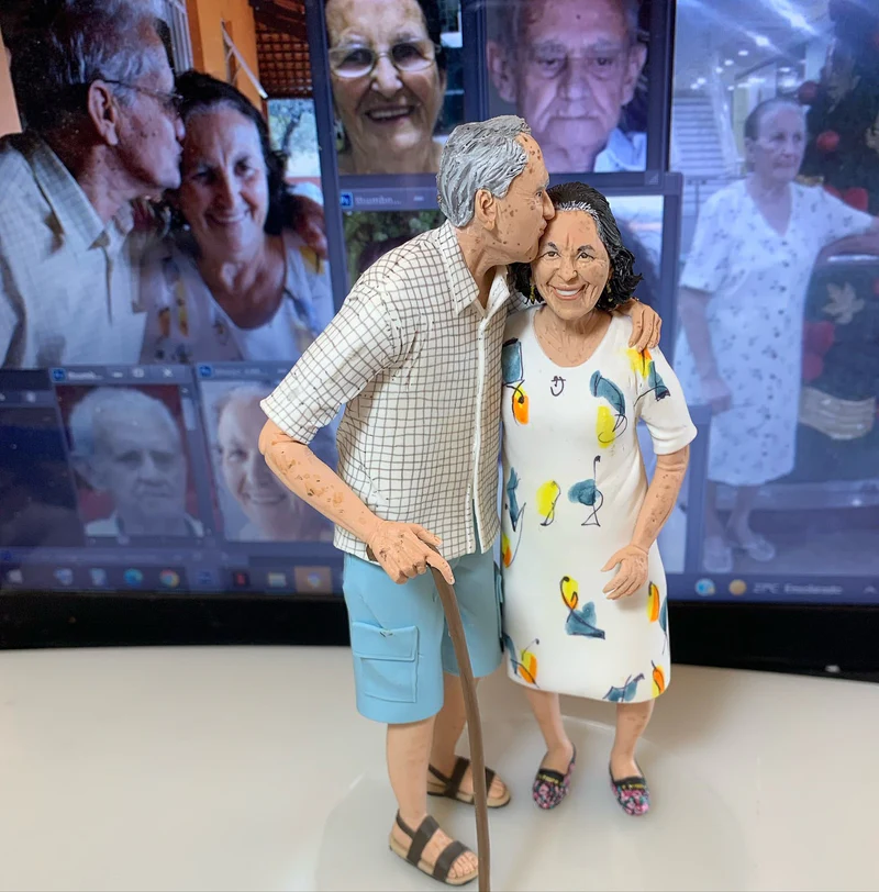 Miniatura esculpida de casal de idosos com o homem beijando a mulher na testa, com a foto original ao fundo