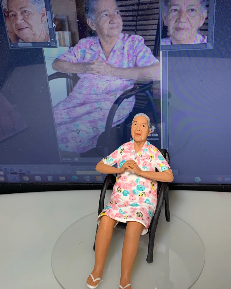 Miniatura esculpida de senhora idosa com vestido florido, com a foto original ao fundo